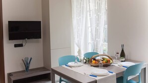 A dining area with a table set for two, a television mounted on the wall, and a window with sheer curtains.