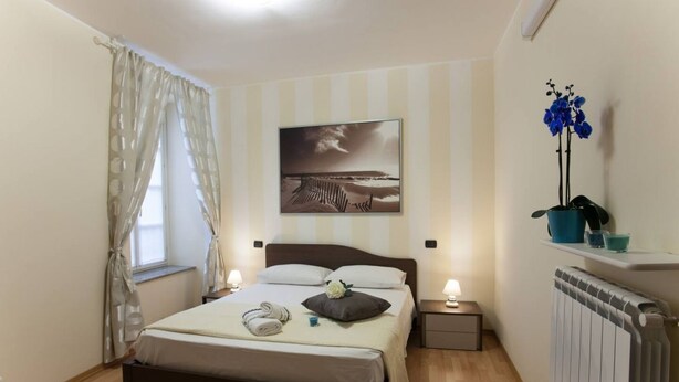 A bedroom with a bed, a nightstand, a window with curtains, a radiator, and a wall-mounted shelf with a plant.
