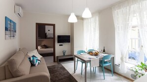 A modern living room with a sofa, a dining table with chairs, a flat-screen TV, and a large window with sheer curtains.