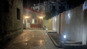 A nighttime view of a courtyard with a tiled floor, a building with a red door, and multiple light fixtures. - A 2018 built, two bedroom, bungalow for short or long term stay (SerreKunda)