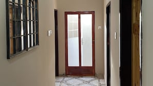 A hallway with a tiled floor, a wooden door, and a window with a decorative frame. - A 2018 built, two bedroom, bungalow for short or long term stay (SerreKunda)