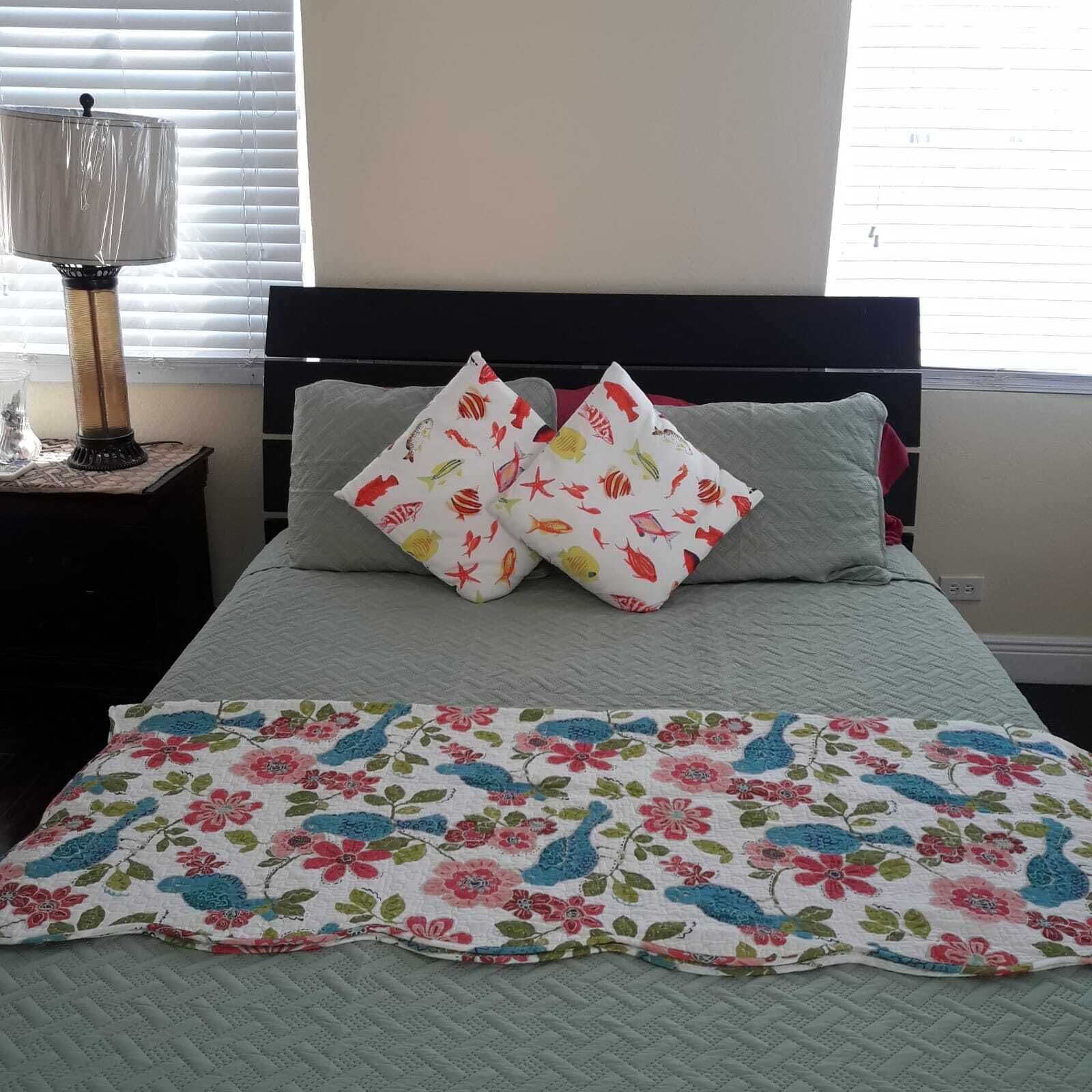 A bedroom with a bed, two pillows with fish patterns, a floral quilt, a lamp, and a window with blinds.