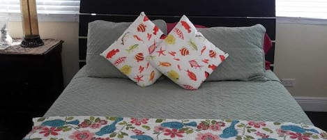 A bedroom with a bed, two pillows with fish patterns, a floral quilt, a lamp, and a window with blinds.