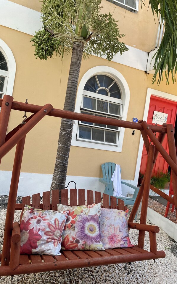 A wooden swing with floral cushions, a palm tree, and a building with a red door and arched windows.