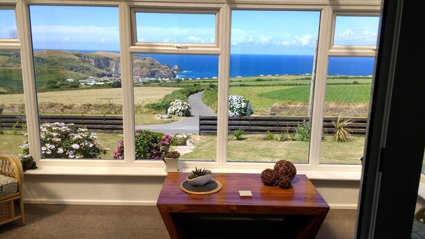 Una habitación con un amplio ventanal que ofrece vistas a un paisaje costero, una mesa de madera con adornos y una silla de mimbre.