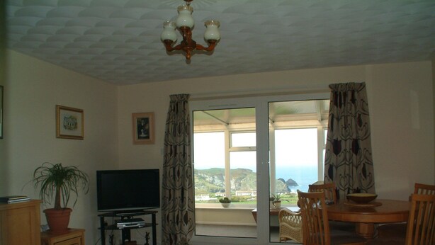 A living room with a television, a dining table with chairs, a potted plant, and a view of the sea through a sliding door. - Luxury Apartment with Spectacular sea views Close to Boscastle/Tintagel (Tintagel, Cornwall)