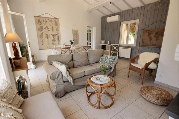 A cozy living room with a sofa, a round wicker table, and a chair. There is a wall-mounted air conditioner and a large plant.