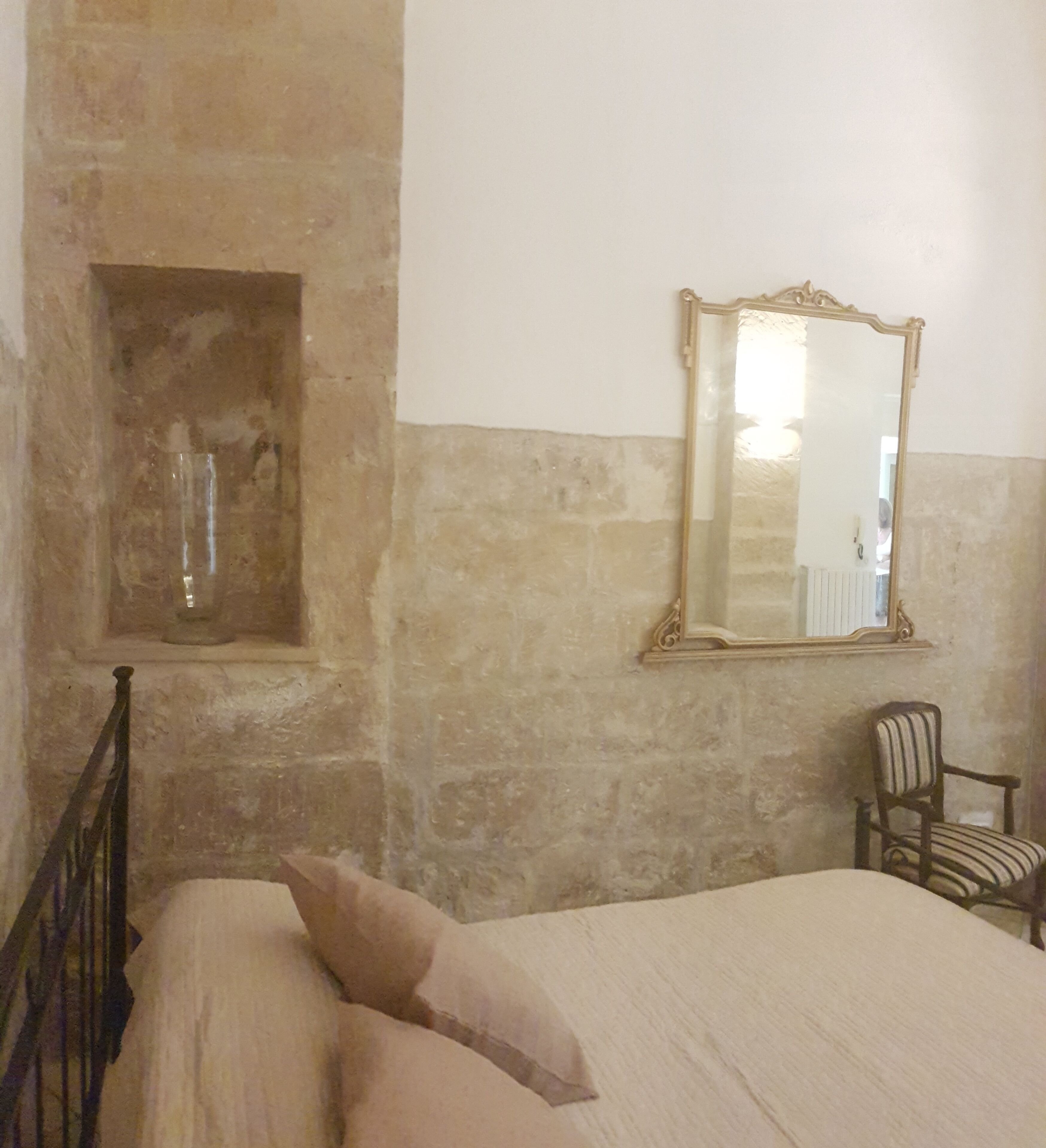 Une pièce comprenant un lit, une chaise, un miroir et une petite alcôve avec un récipient en verre.
