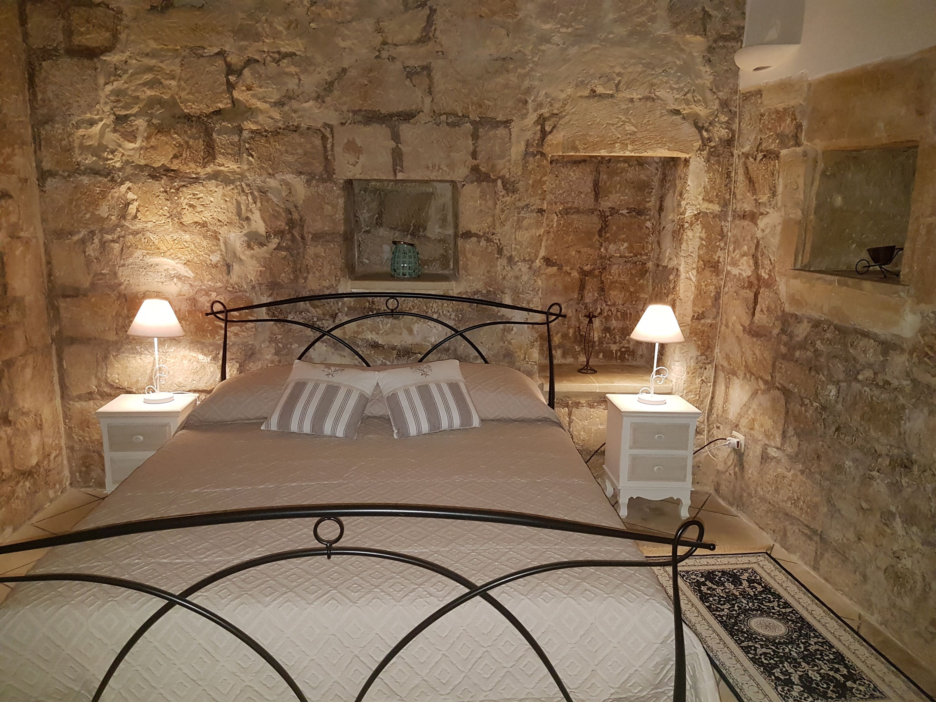 Ein Schlafzimmer mit Steinwand, einem Bett mit Metallgestell, zwei Nachttischen mit Lampen und einem kleinen Regal mit einer Dekoration.