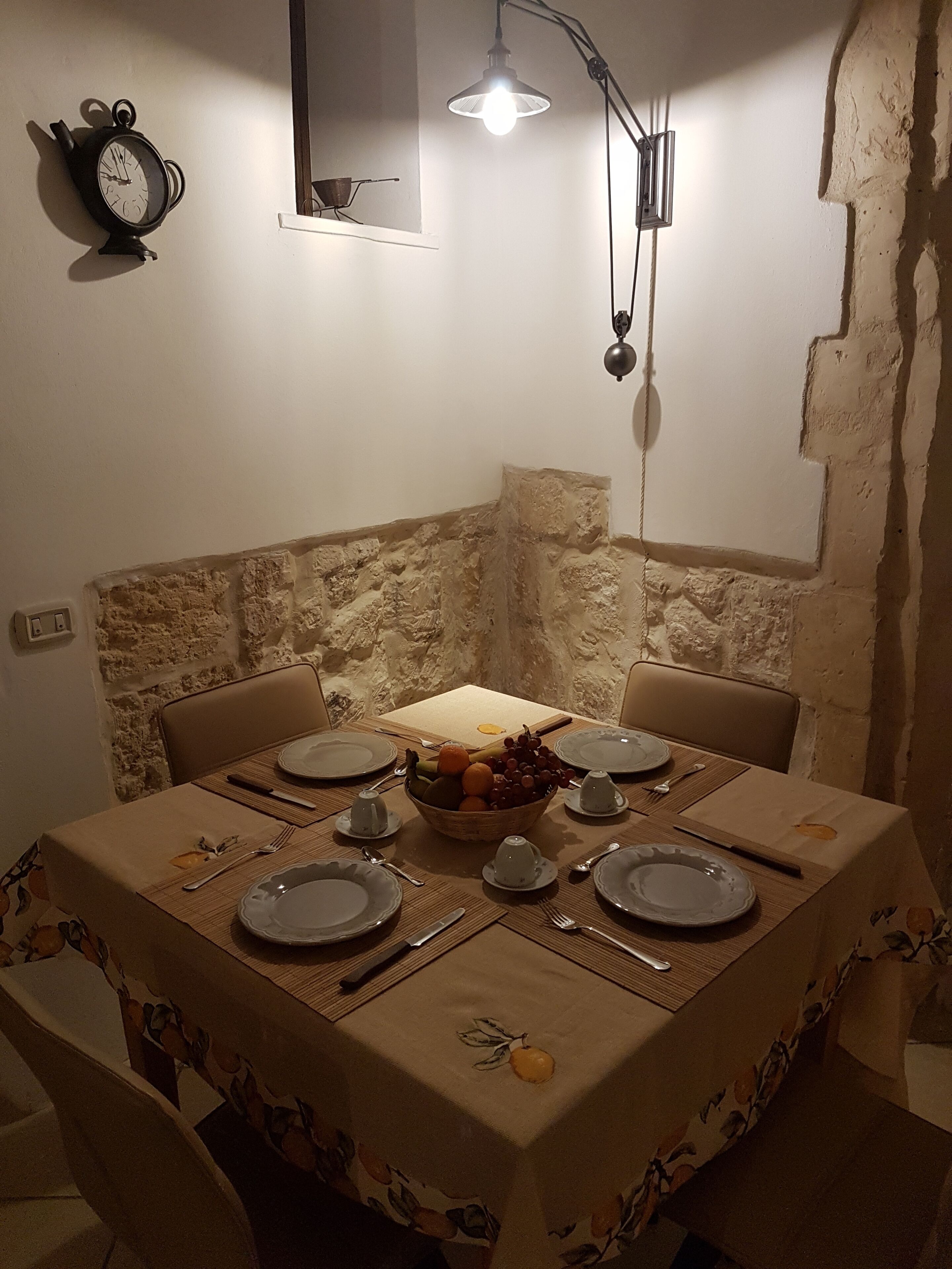 Un coin repas avec une table dressée pour quatre personnes, un panier de fruits et une horloge au mur.