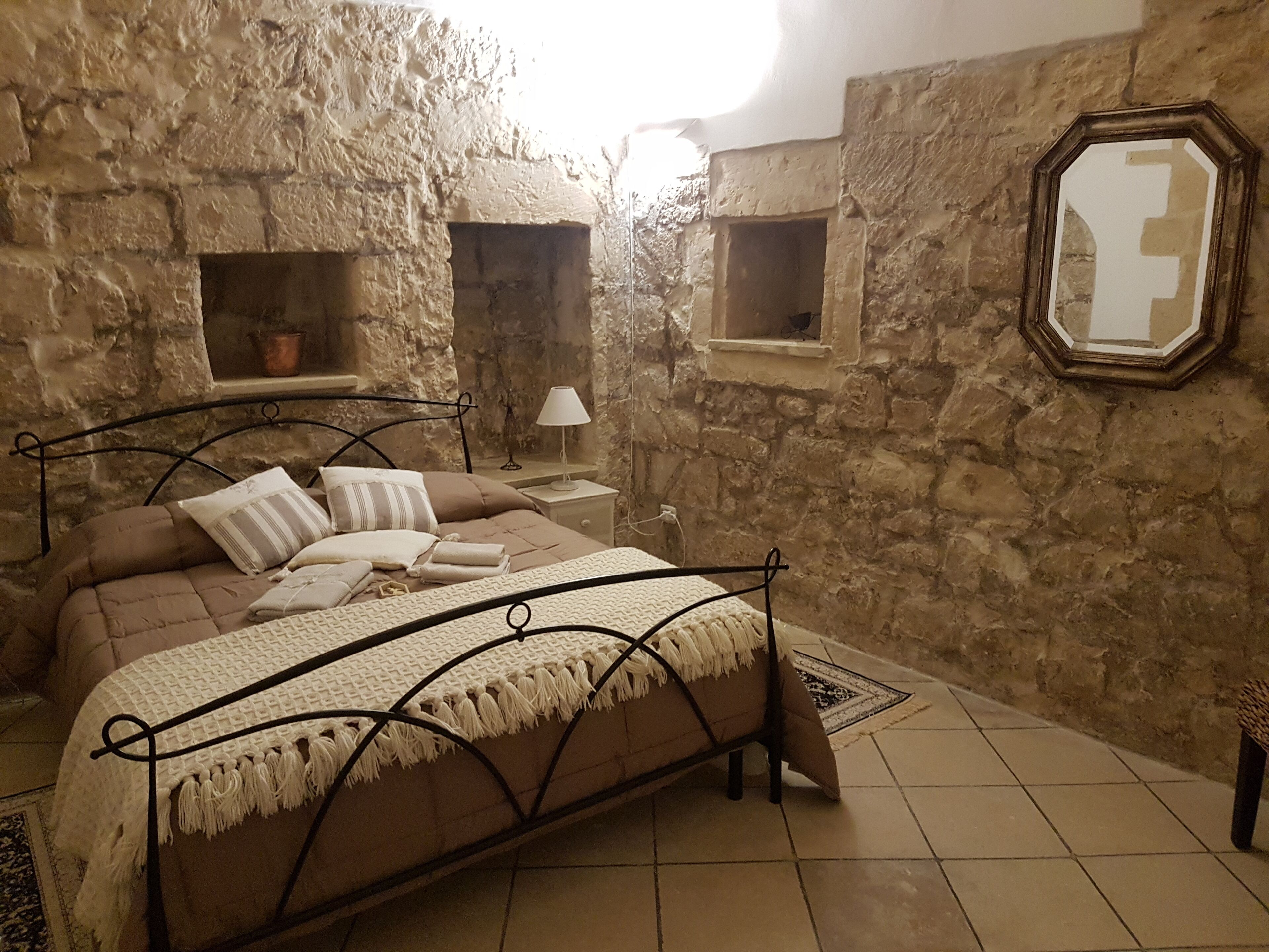 A bedroom with a stone wall, a bed with a metal frame, a nightstand, and a mirror.