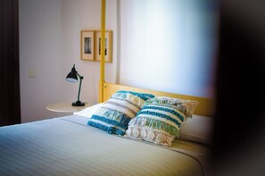 Ein ordentlich bezogenes Bett mit gestreiften Kissen, ein kleiner Nachttisch mit einer Lampe und ein gerahmtes Bild an der Wand.
