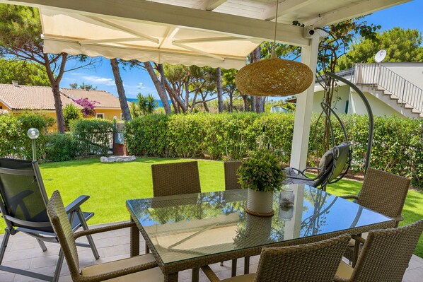 An outdoor patio with a glass table and wicker chairs, surrounded by greenery and a view of a residential area. - Sunset Holiday Villa in San Pietro Pinewood (Valledoria)