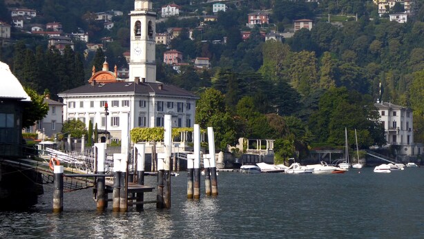 A waterfront building with a clock tower, docked boats, and a backdrop of hillside residences. - Lovely house close to the lake with panoramic lake and mountain views (Acquaseria, San Siro)