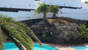 A pool area with two swimming pools, palm trees, and a rocky wall. - Front line, contemporary,  refurbished, stunning sea views, pool, WiFi, SKY (Puerto del carmen)