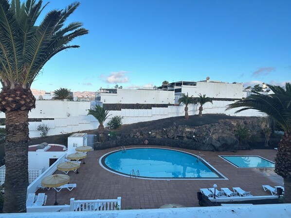 A pool area with lounge chairs and umbrellas, surrounded by palm trees and a white building with satellite dishes. - Front line, contemporary, refurbished, stunning sea views, pool, WiFi, SKY (Puerto del carmen)
