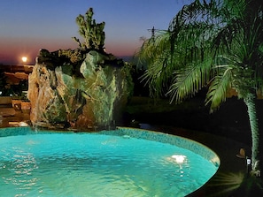 Une piscine nocturne agrémentée d’une cascade artificielle en roches, entourée de palmiers et d’un mur de pierre.