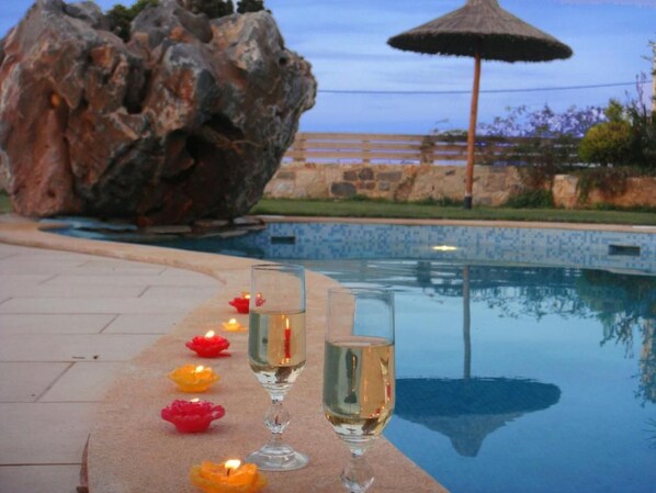 A poolside area with a large rock formation, a thatched umbrella, and candles on the edge of the pool.