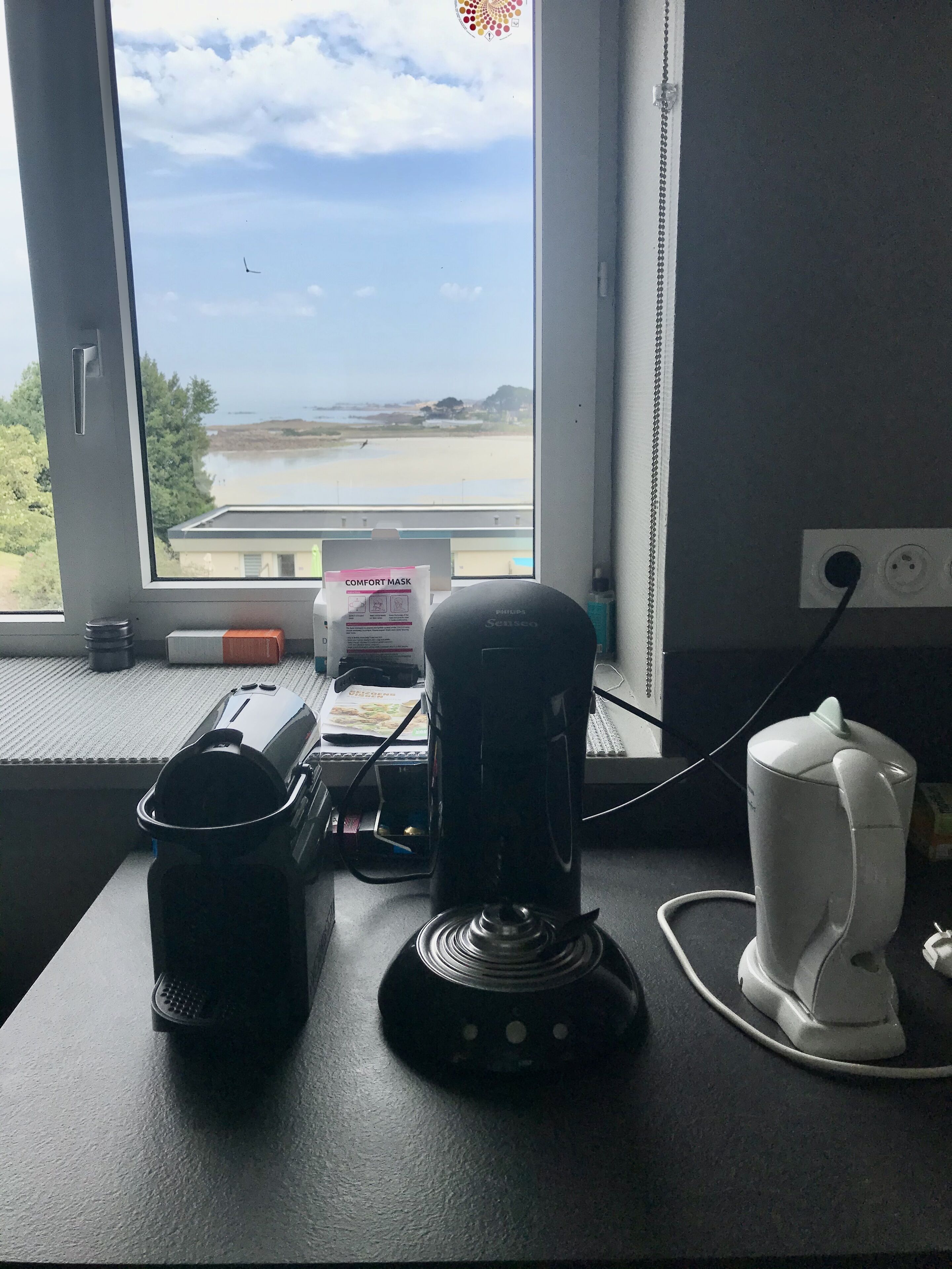 Un bureau avec une machine à café, une tasse blanche et une boîte étiquetée « MASQUE CONFORT », près d’une fenêtre donnant sur une plage.