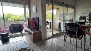 Uma sala de estar com televisão, um espaço de jantar com mesa e cadeiras, e uma varanda com vista para árvores.