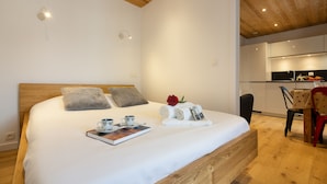 Een net opgemaakt bed met een boek, een beker en handdoeken, een houten hoofdbord en een kleine kitchenette op de achtergrond.