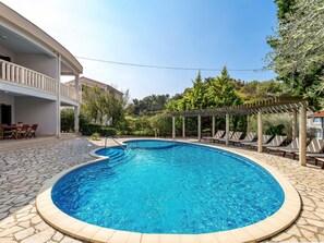 Una piscina circular con solárium de piedra, un área de estar cubierta con sillas y un edificio con balcón al fondo.