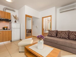 Una sala de estar moderna con sofá, reposapiés y mesa de centro, una cocina con refrigerador y estufa, y un espejo grande que refleja la habitación.