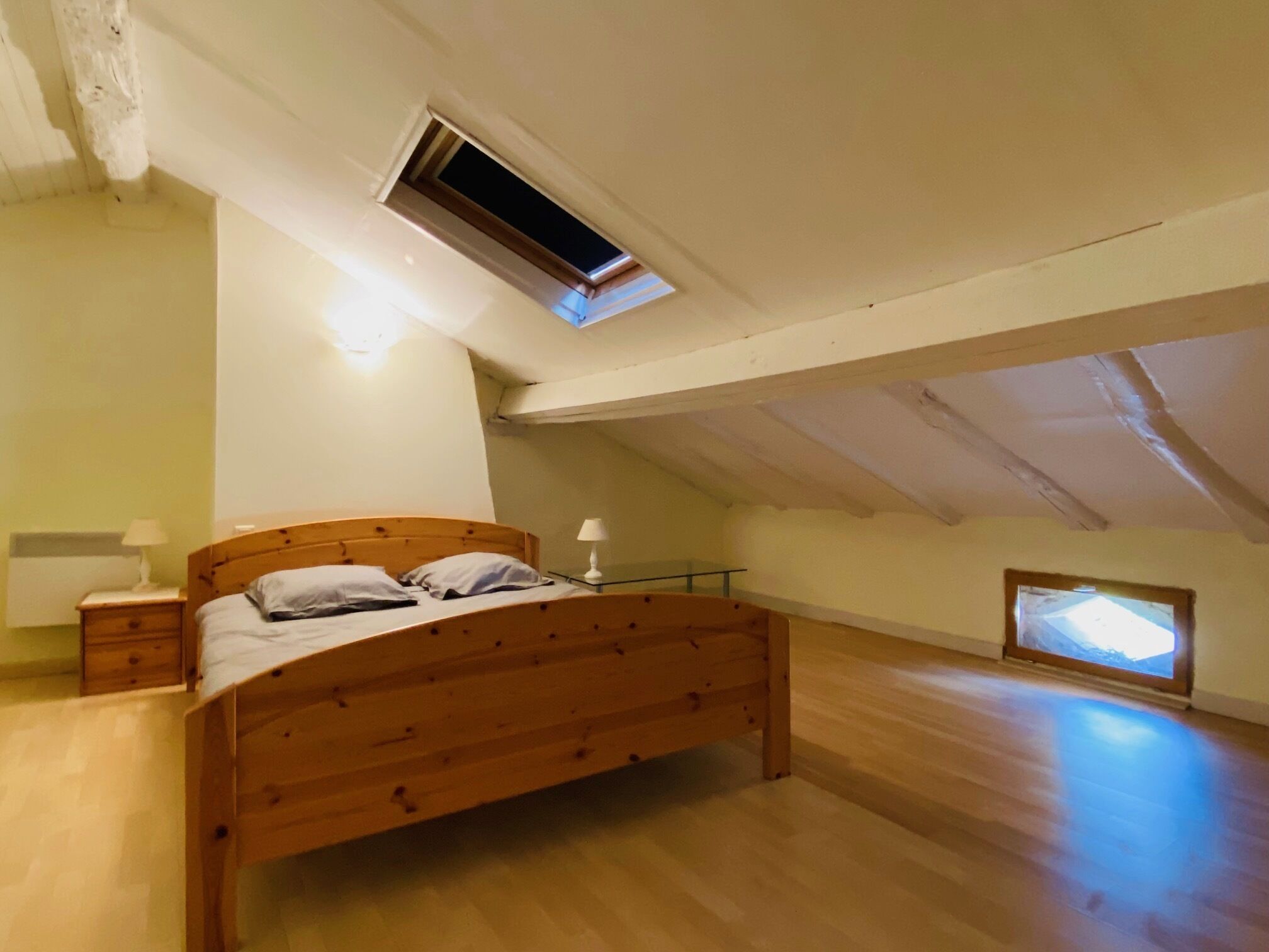 A bedroom with a wooden bed, a nightstand, a lamp, a small window, and an attic space with a skylight.