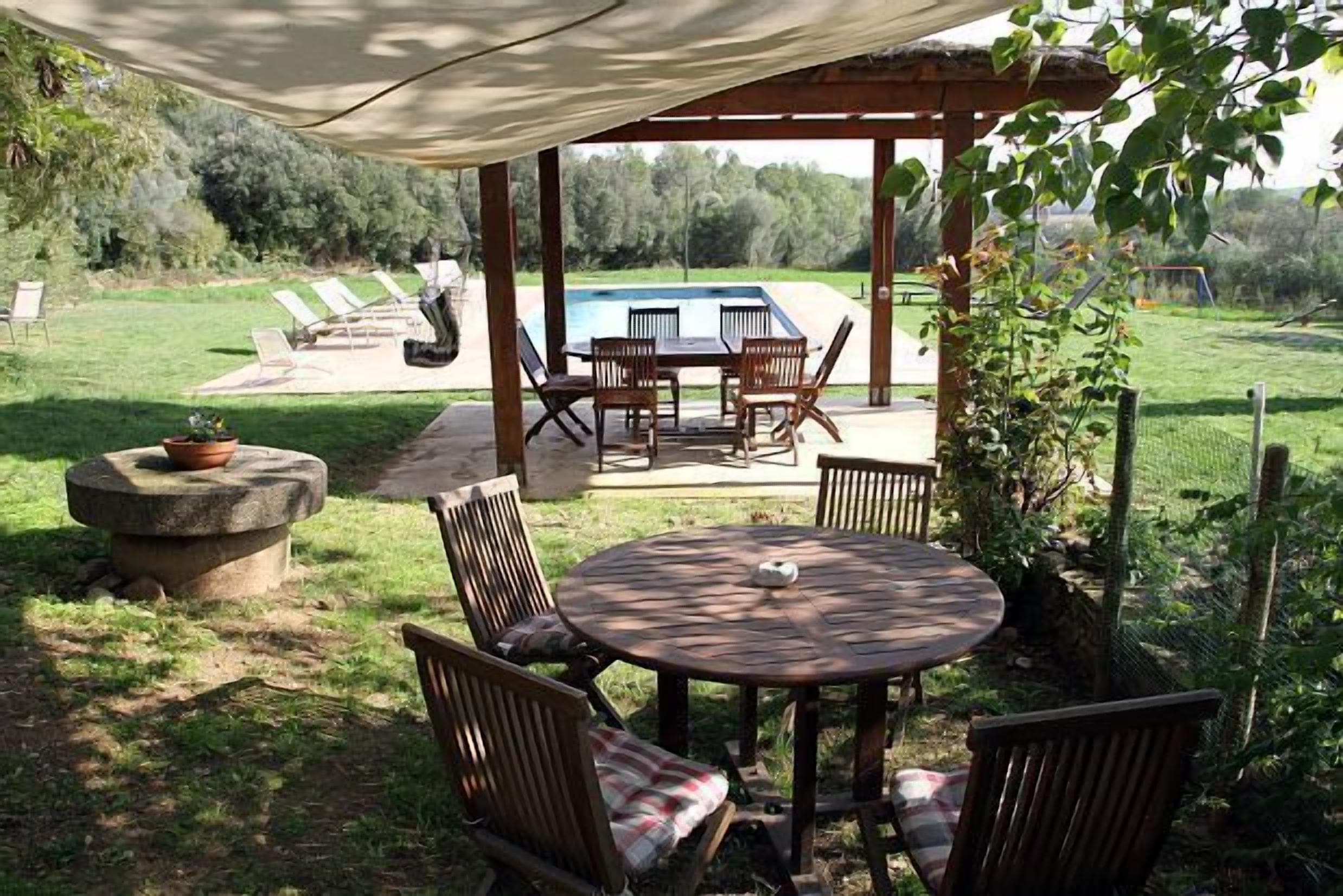 An outdoor patio with a round table and chairs, a stone planter, and a view of a swimming pool and lawn.