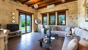 A spacious living room with stone walls, a wooden ceiling, a large TV, a sofa, and a coffee table with a vase and wine glasses.
