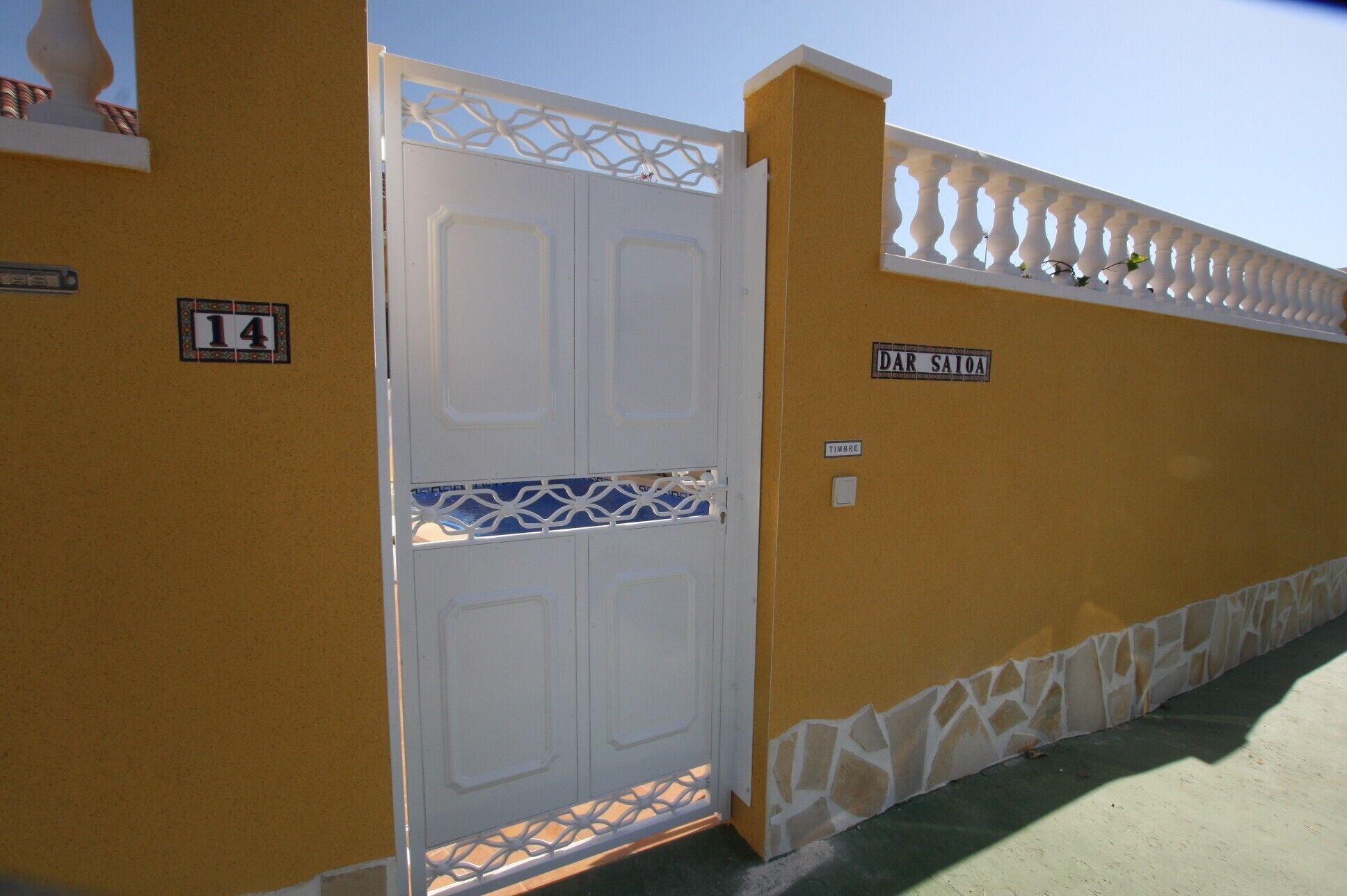 Un portail blanc orné d’un treillis métallique décoratif, un mur jaune avec un socle en pierre, et une enseigne indiquant « DAR SAIOA ».