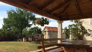 An outdoor patio area with a wooden table and bench, surrounded by stone walls and greenery.