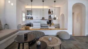 A modern kitchen with white cabinets, a central island, hanging pendant lights, and a small dining area with a round table and ottoman.
