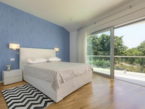 Quarto com uma cama grande, mesa de cabeceira, um vaso e vista para o verde através de uma porta de correr de vidro.