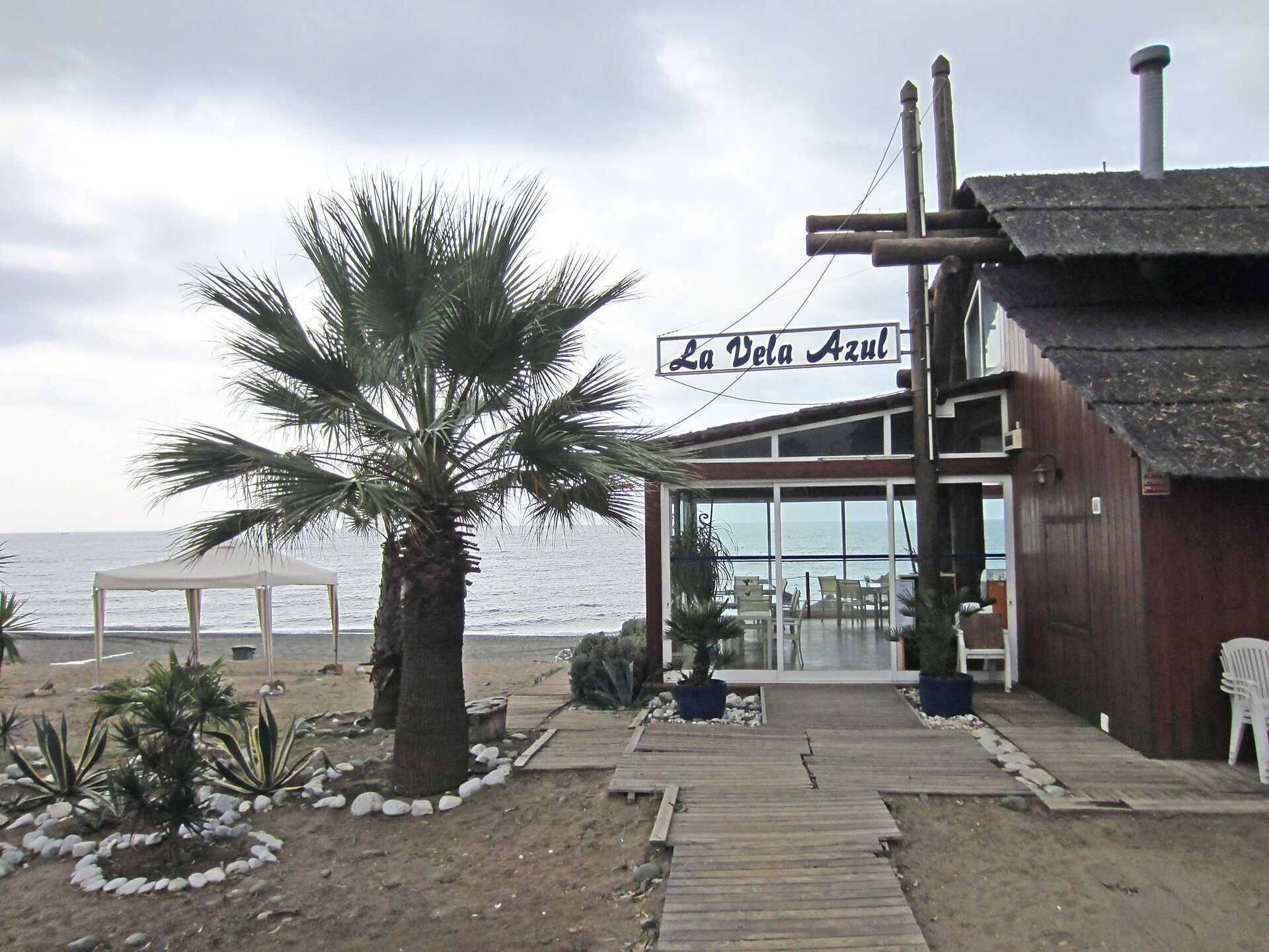 Un edificio frente a la playa llamado "La Vela Azul" con una terraza de madera que conduce a un área cubierta con sillas y mesas, con vista al mar.
