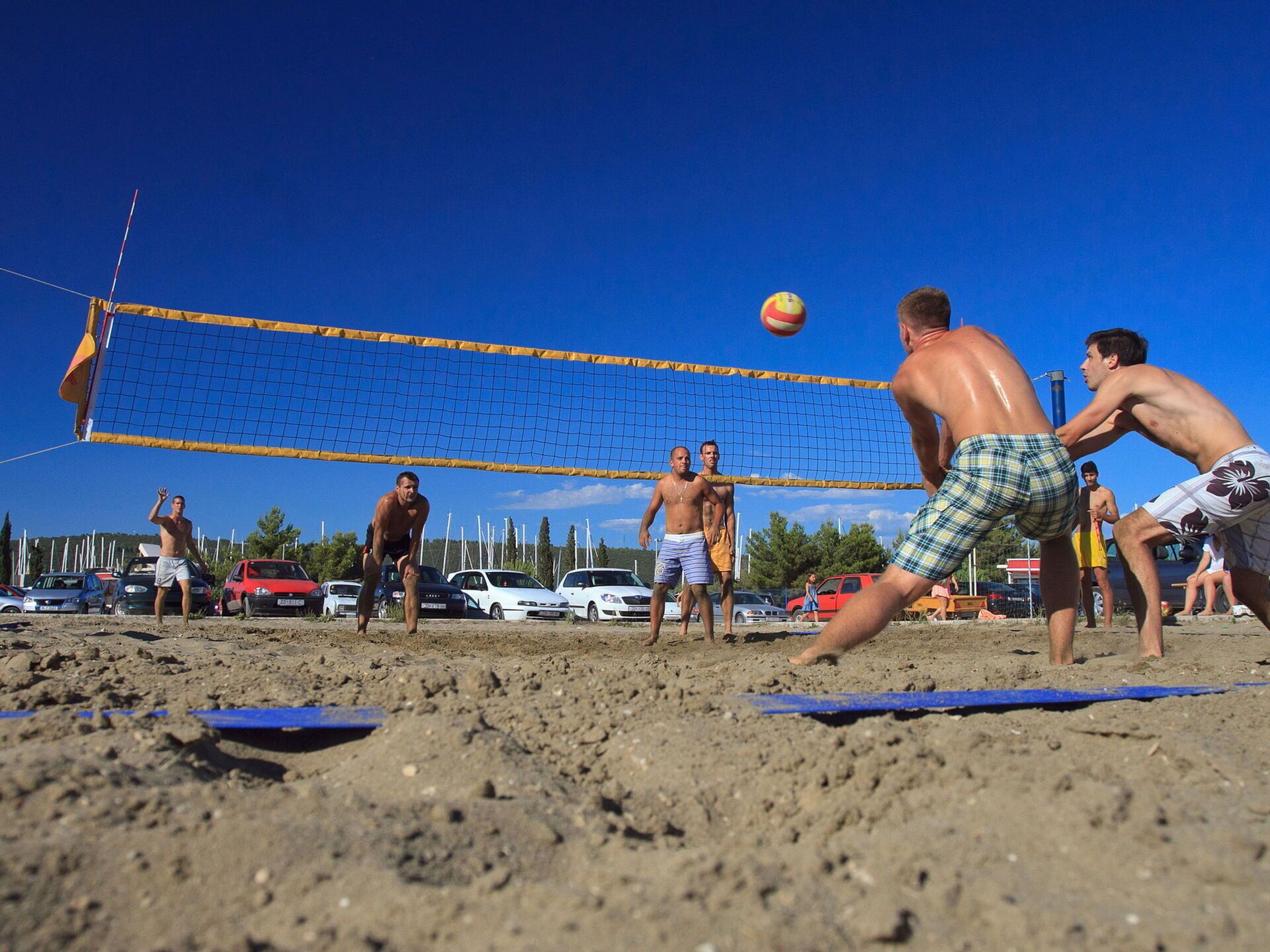 Een strandvolleybalwedstrijd met spelers in korte broeken zonder shirt, een blauw net en auto's op de achtergrond.