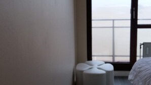 Quarto de hotel com uma cama, um banquinho pequeno branco, uma janela com vista e uma porta com sinal de saída de emergência.