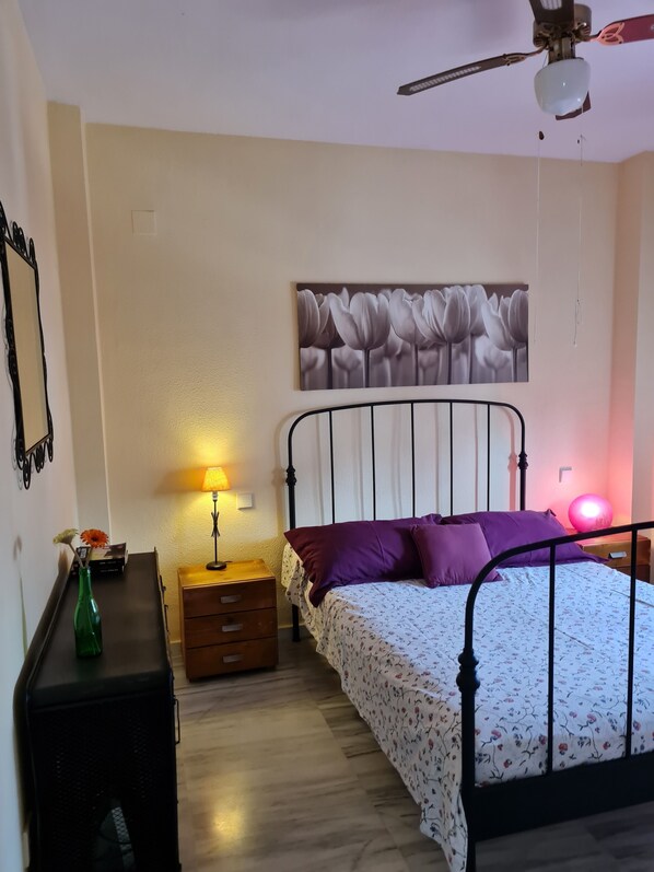 A bedroom with a bed, a nightstand, a lamp, a mirror, and a ceiling fan.