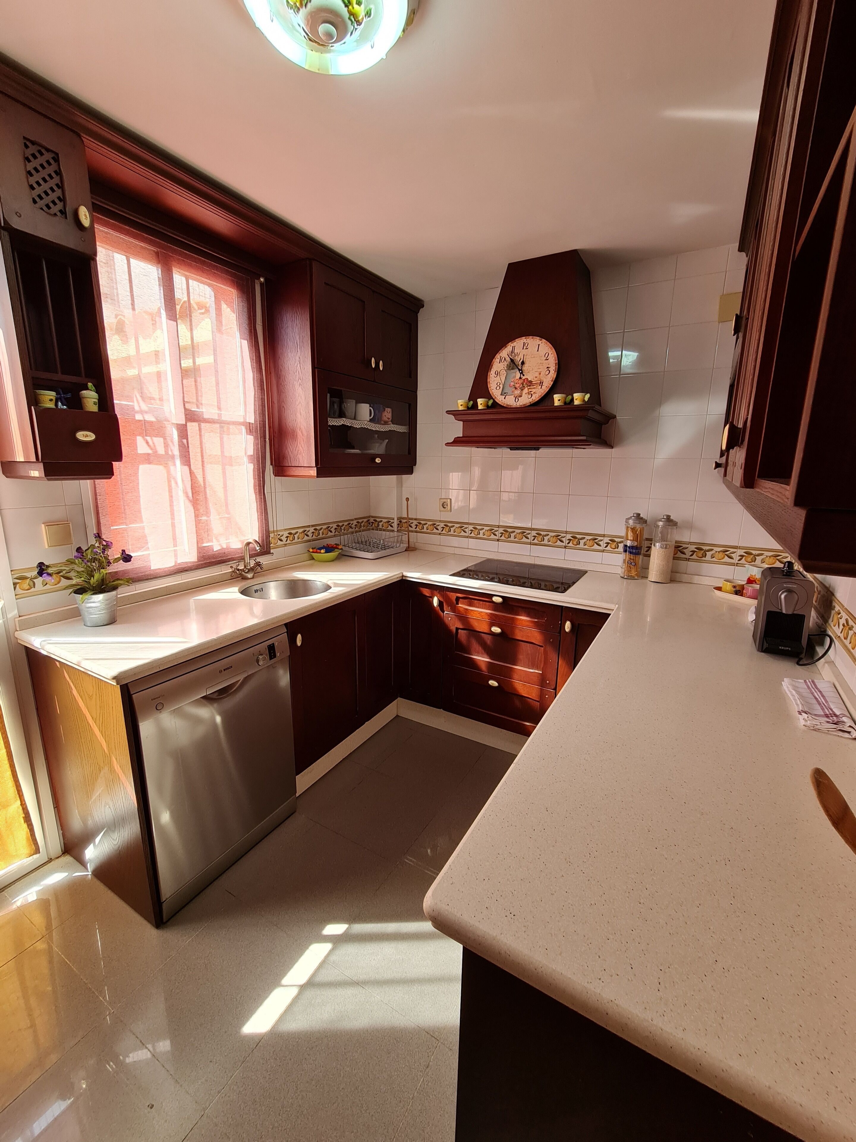 Een keuken met houten kastjes, een roestvrijstalen vaatwasser, een spoelbak en een klok aan de muur.