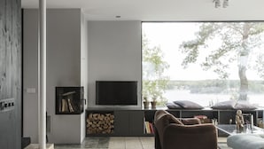 A modern living room with a large window, a flat-screen TV, a sofa, and a staircase leading to another level. - Exclusive architectural house in archipelago (Stockholm)