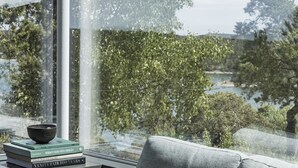 A modern living room with a large window, a grey sofa, and a small table with books and a bowl. - Exclusive architectural house in archipelago (Stockholm)