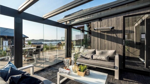 Een moderne buitenruimte met een glazen omheining, een houten terras en een zithoek met kussens en een tafel.