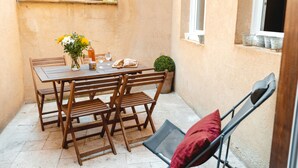 A patio with a wooden table set for two, a small potted plant, and a window with a view of yellow flowers.