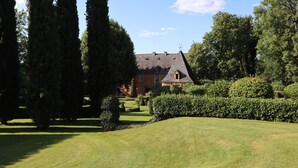 A large, historic building with a steeply pitched roof and multiple chimneys, surrounded by well-maintained hedges and tall trees.