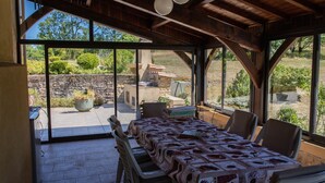 Een terras met eettafel gedekt voor vier personen, omringd door stoelen, onder een houten pergola met grote ramen die uitzicht bieden op de tuin en de buitenkeuken.