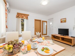 Una sala de estar moderna con comedor, que incluye un televisor de pantalla plana, un mueble de televisión de madera, un mantel floral y una mesa lista para el desayuno con frutas y jugo.