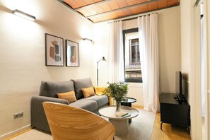 Ett modernt vardagsrum med en grå soffa, en trästol, ett runt soffbord med en bok och en växt, ett TV-bord och ett fönster med gardiner.