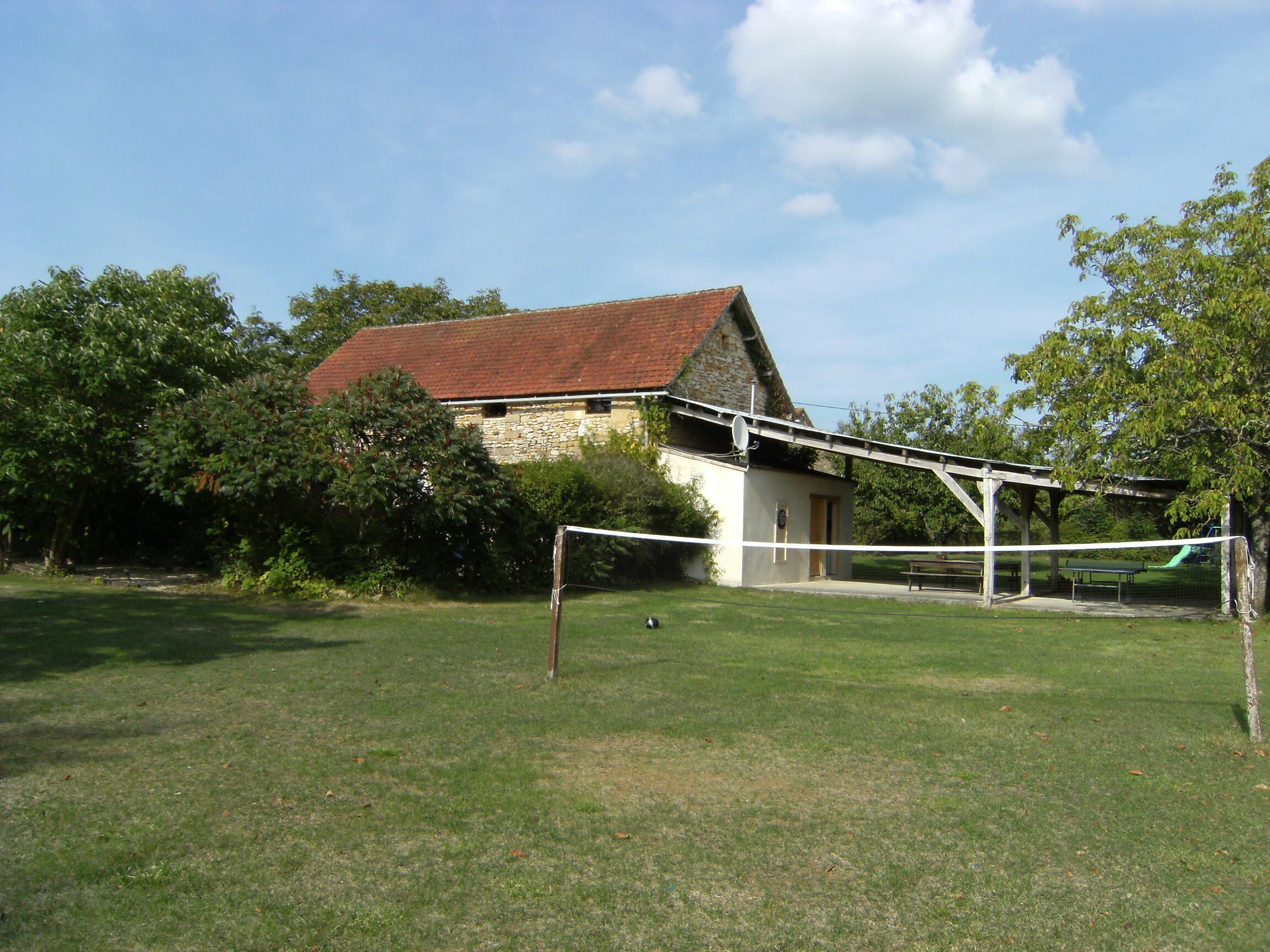 A grassy area with a volleyball net, a stone building with a red roof, and a bench.