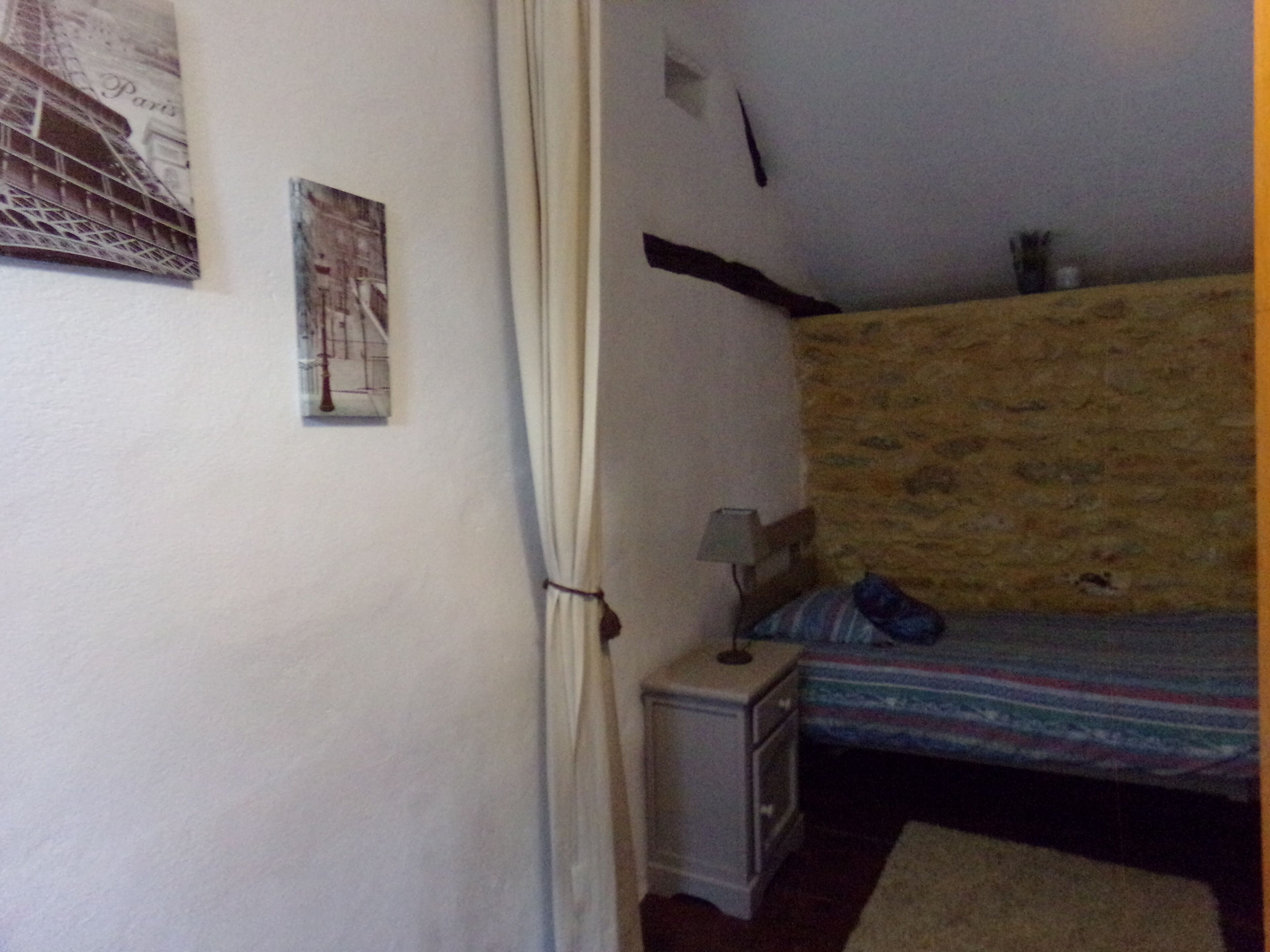 Un dormitorio con cama, mesita de noche, lámpara, estante en la pared y ventana con cortinas.