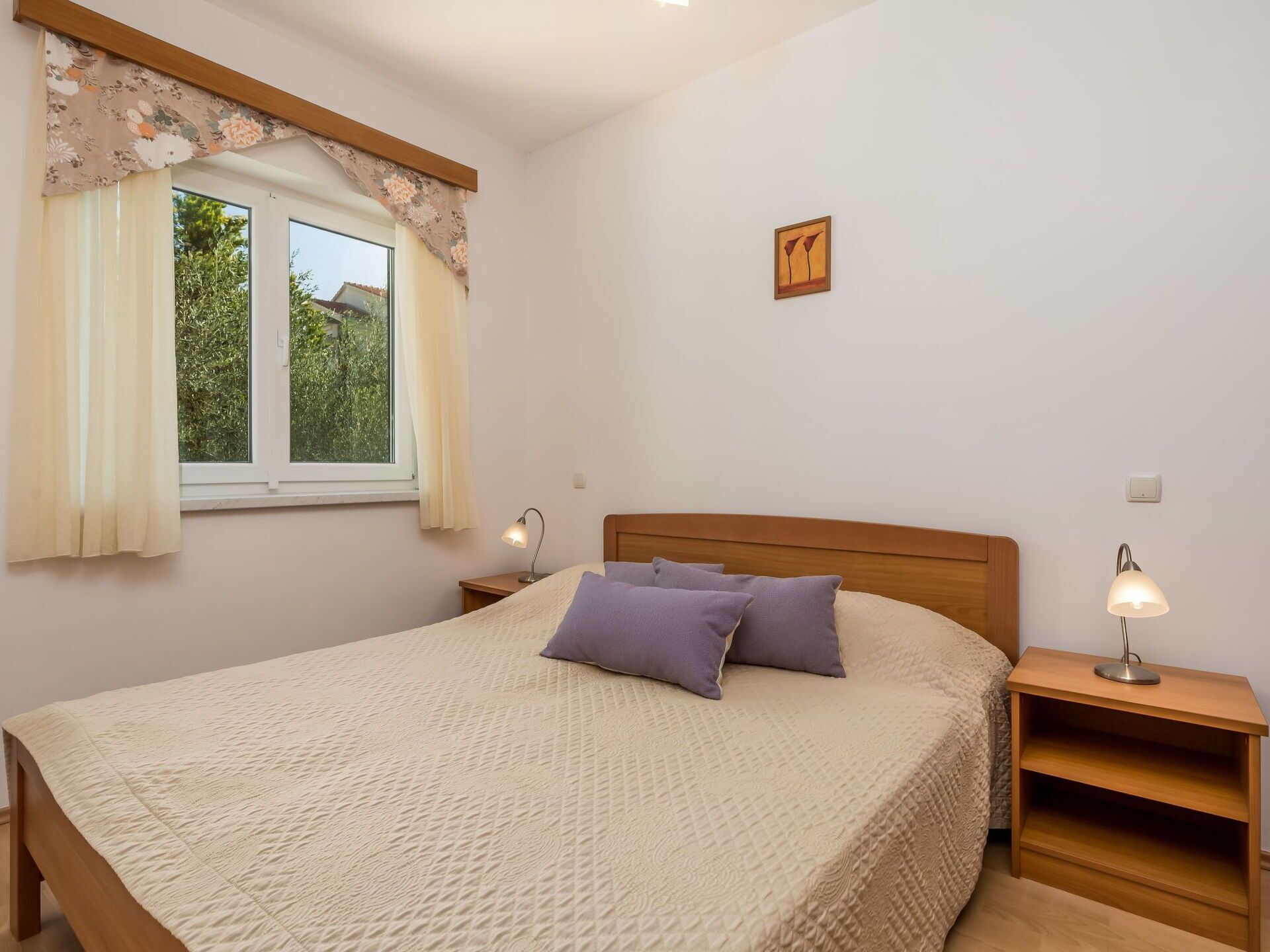 A bedroom with a bed, two bedside tables, a window with curtains, a wall-mounted picture, and a nightstand with a lamp.